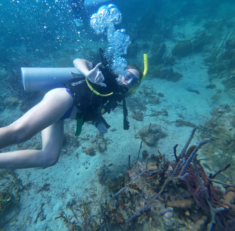 The image shows a woman scuba diving in clear blue water. She is wearing a black wetsuit, a yellow snorkel, and a mask. She has a tank on her back and is giving a hand signal. The seabed is visible with sand and some coral formations. Bubbles are rising towards the surface, indicating she is exhaling. The overall scene suggests an underwater exploration or recreational diving activity.